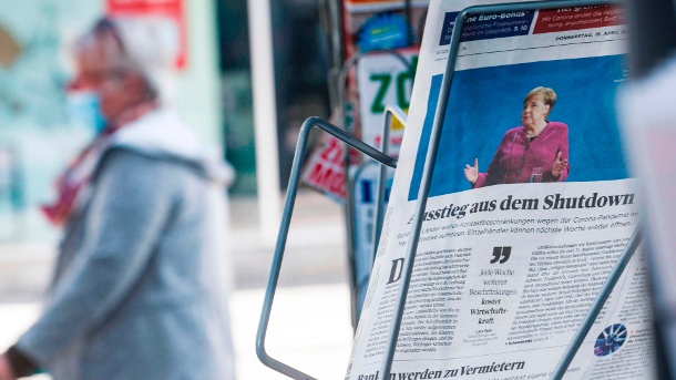 Eine Frau mit Mundschutz geht an einem Zeitungsständer vorbei: Auf der Titelseite einer Wirtschaftszeitung steht unter einem Bild von Bundeskanzlerin Merkel 